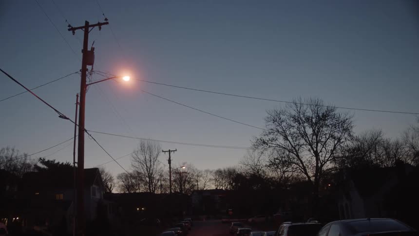 A street light is lit on a neighborhood street with cars parked on both sides
