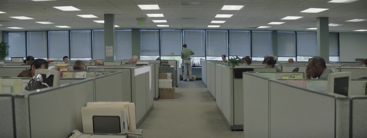 A man walks past cubicles to pour a cup of coffee and look out the window