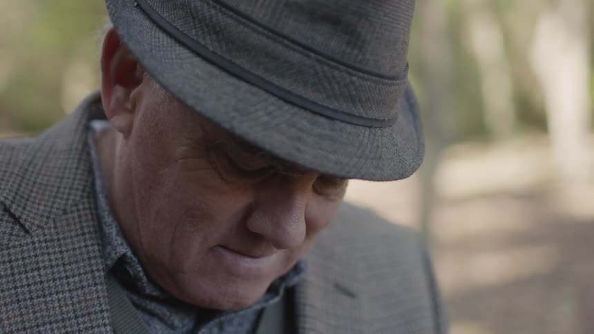 An older man in a hat grimaces and shakes his head