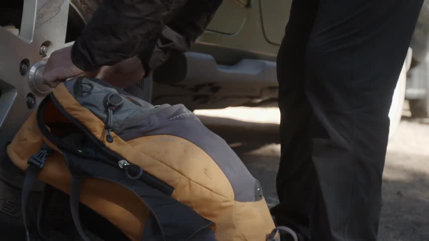 A man zips up a backpack that is on the ground