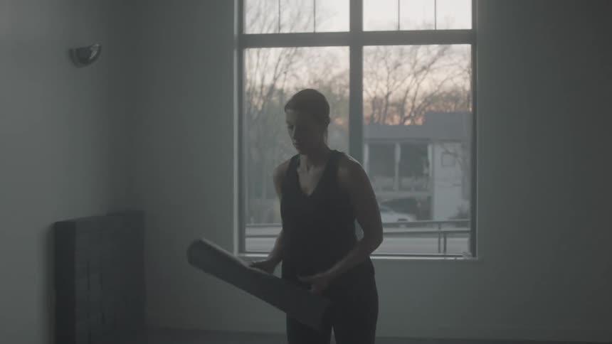 A woman holding a yoga mat walks toward a window