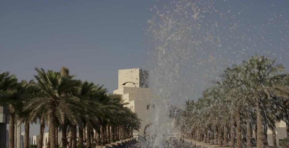 A water fountain sprays in front of a building and palm trees