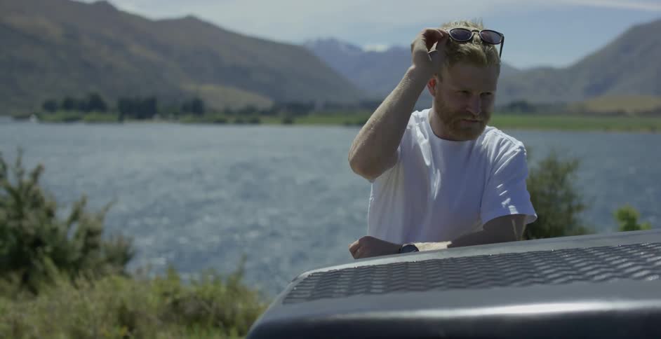 A man leaning on a car puts his sunglasses on his head