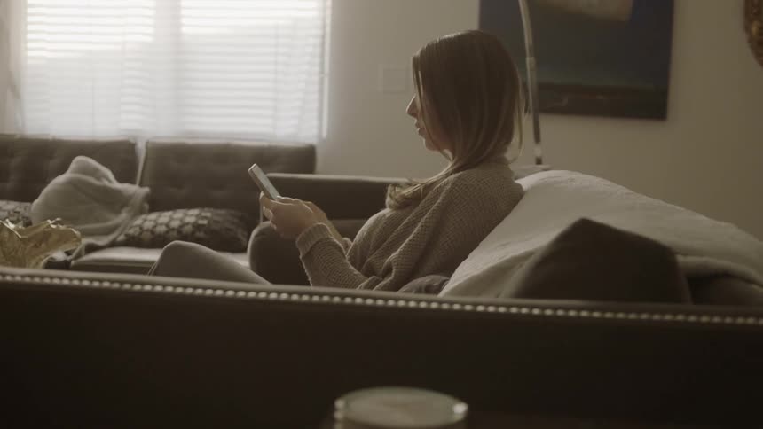 A woman sits on her couch scrolling on her phone
