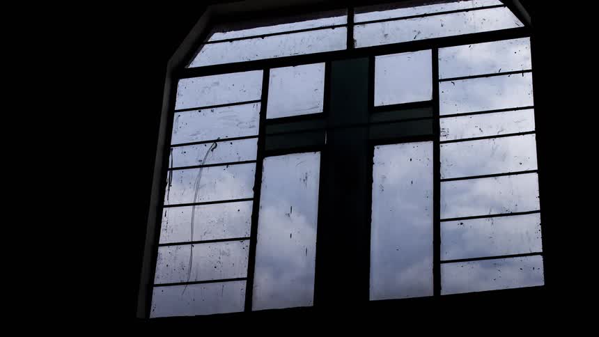 Storm clouds move through the sky beyond a window with a cross
