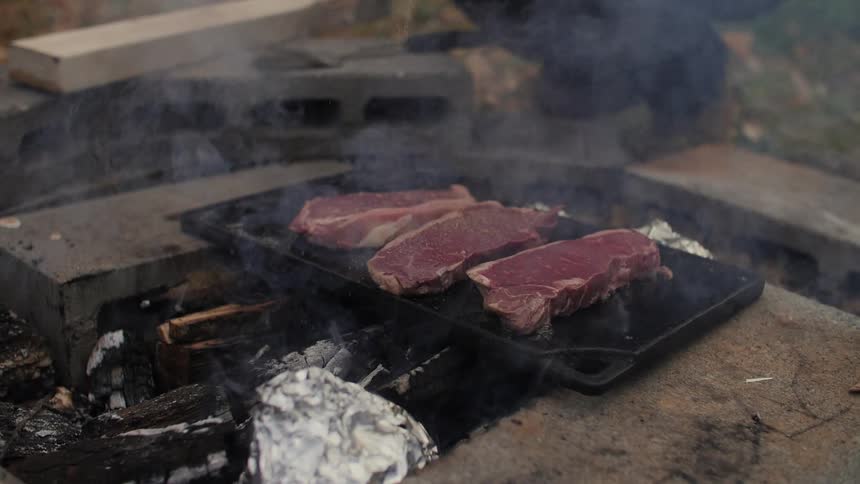 Steaks cook on an outdoor fire