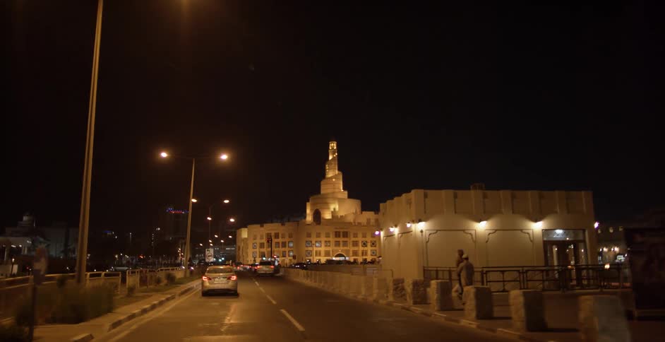 Cars drive on a street toward a well lit building at night