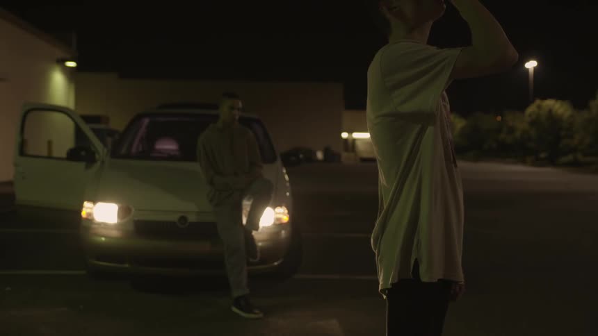 Two men in a parking lot next to a parked car at night