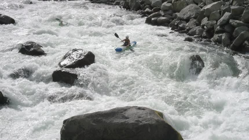 Two white people in kayaks ride through a massive river rapids