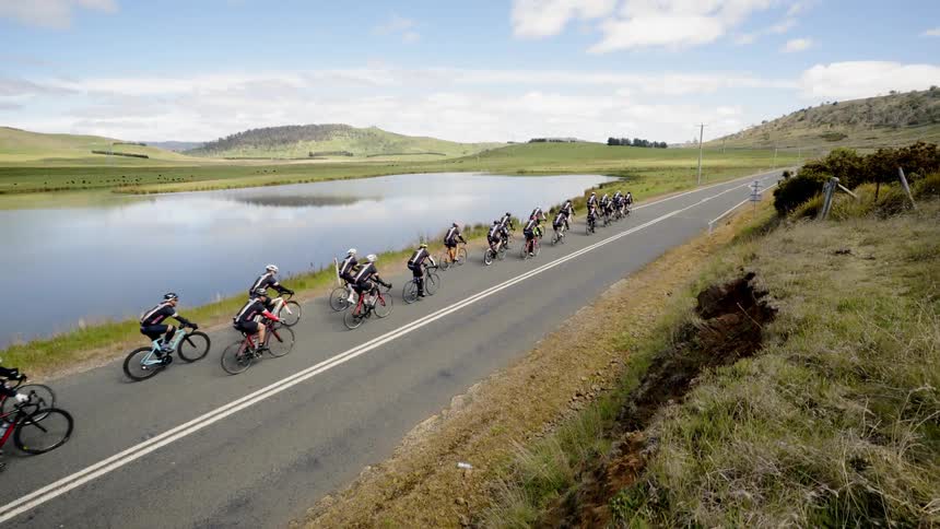 A group of bicyclists in a peloton travel on a country road