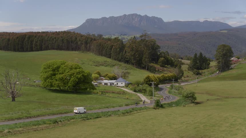 Bicyclists ride on a path in the country by a house and hills