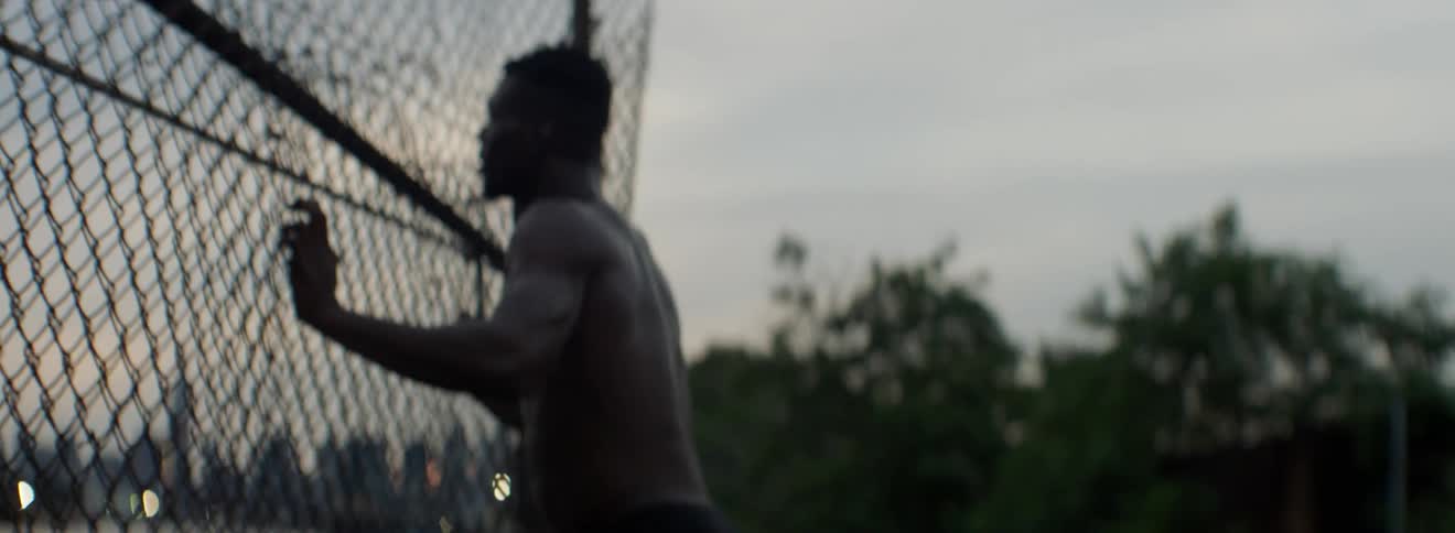 A shirtless man stands next to a chain link fence at sunset