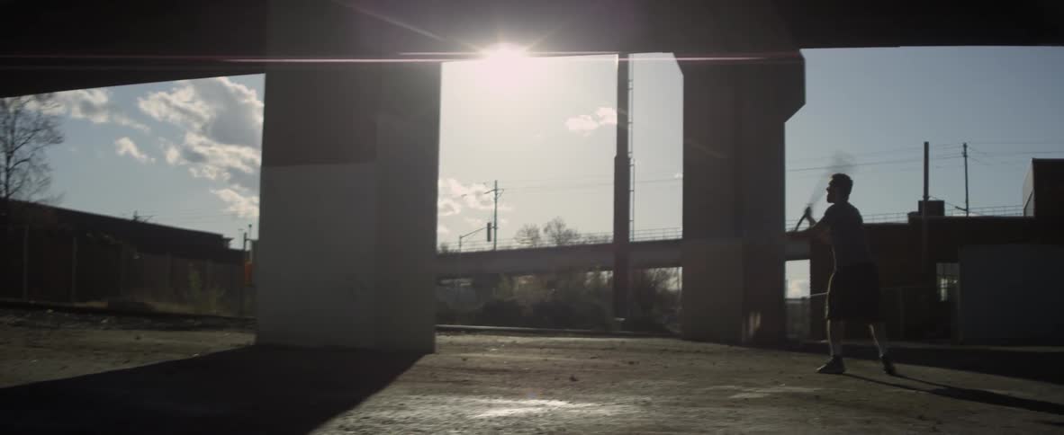 A man bounces a ball beneath a bridge and catches it with a lacrosse stick
