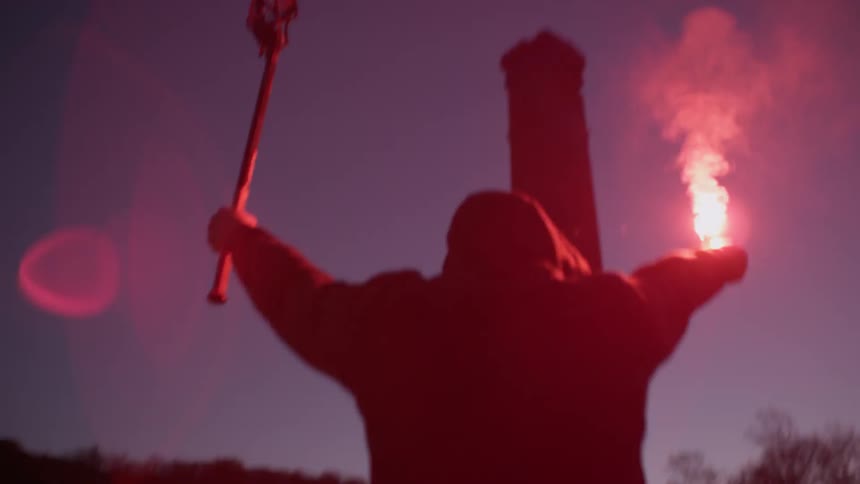 A man is raising a lacrosse stick and flare into the dawn sky