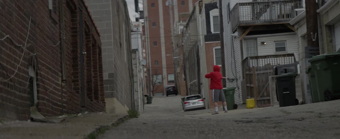 Man shooting ball from lacrosse net against brick building wall in alley
