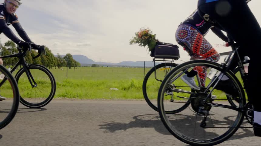 People ride bicycles on a road passing a farm field
