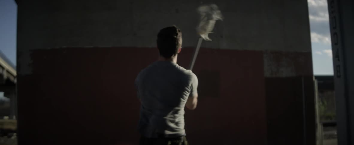 A man bounces a ball off a wall and catches it with a lacrosse stick