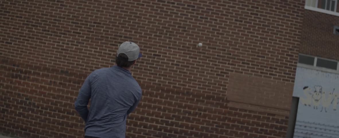 A man bounces a ball off a building and catches it with a lacrosse stick