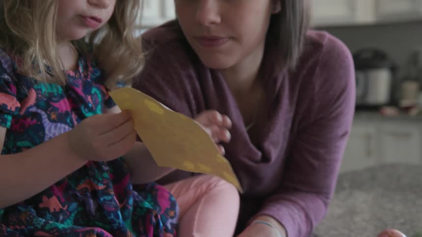 Child sitting on a kitchen counter looks with her mom at a sheet of stickers to decorate easter eggs
