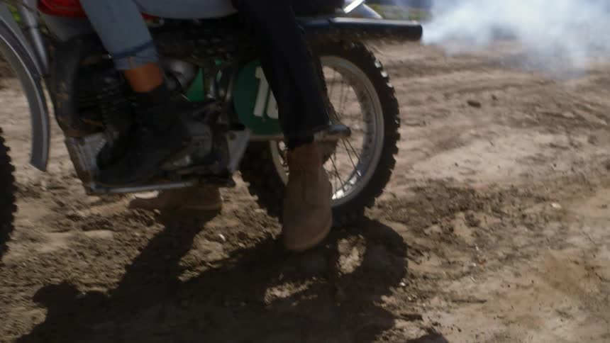Two people sitting on a motorbike and one starts it with their foot