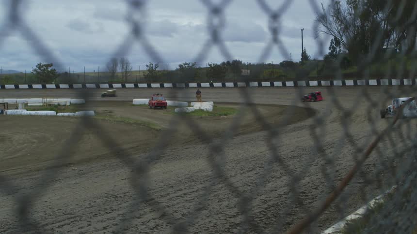 Race cars running around a dirt track as seen from just outside a fence on a cloudy day