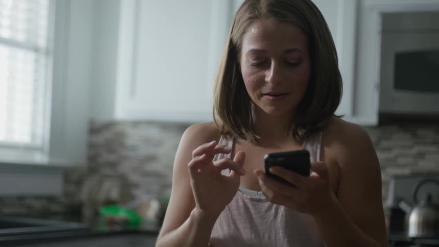 A woman standing in a kitchen is looking at her cellphone