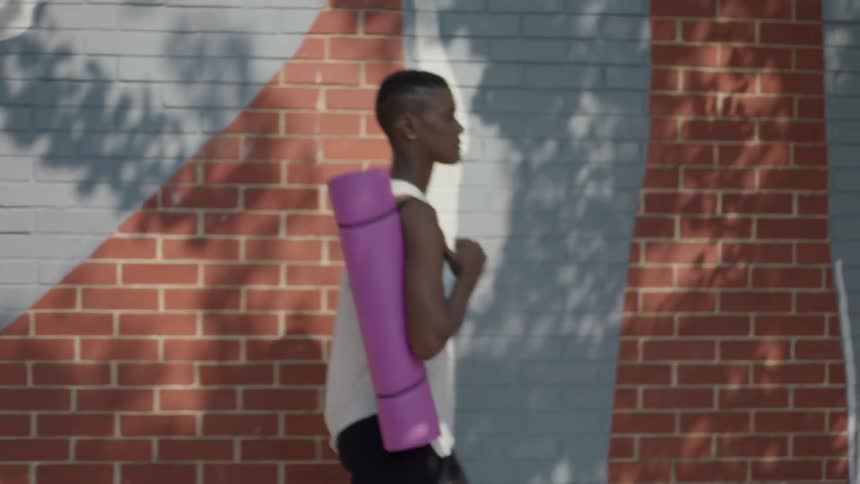 A black person carrying a pink exercise mat walks next to a brick building