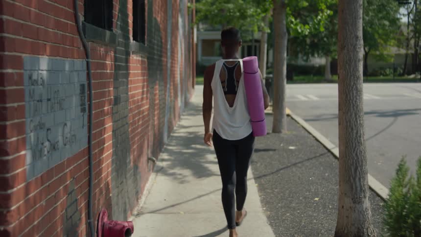 A black woman carrying a yoga mat is walking past a brick wall
