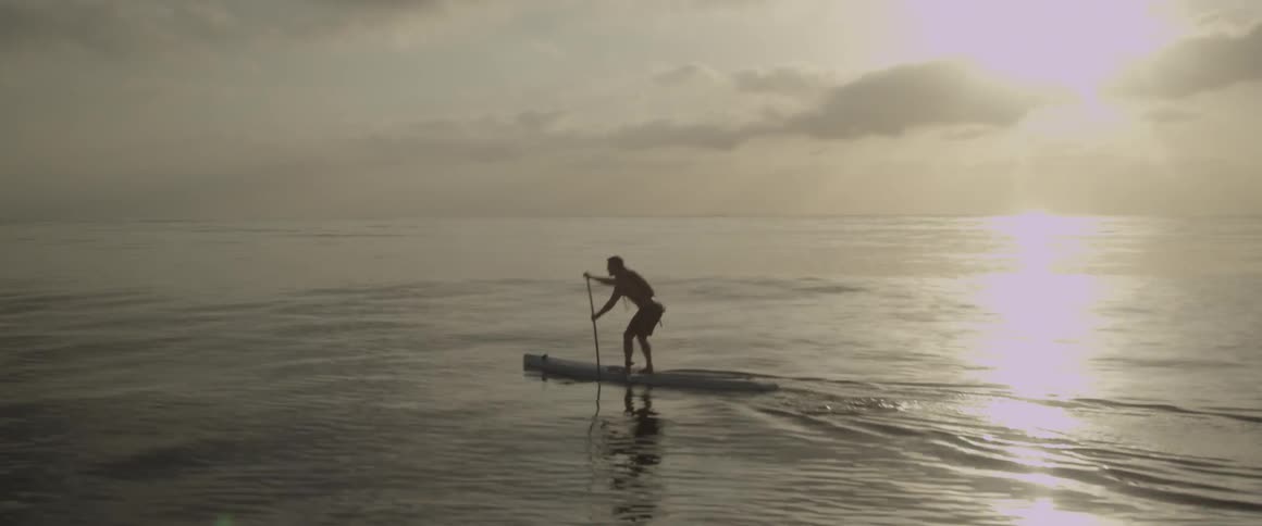 A man stands on a kayak as he paddles through the ocean under a sunset