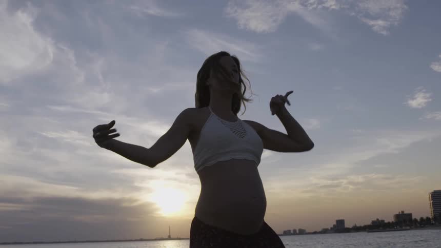 A pregnant woman dances near the ocean at sunset