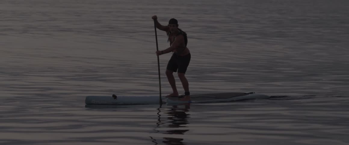 A man standing on a kayak paddles through the ocean