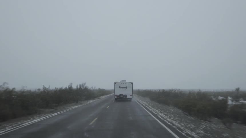 A trailer is driving down a two lane road while it is snowing