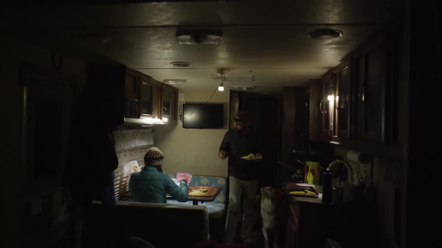 A man and woman with a dog have dinner inside a camper trailer