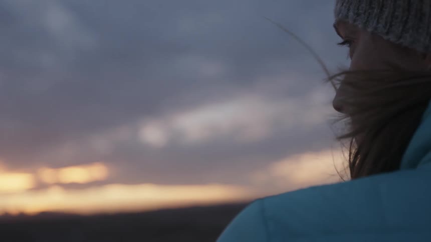 A woman is standing outside on a windy day watching the sunset