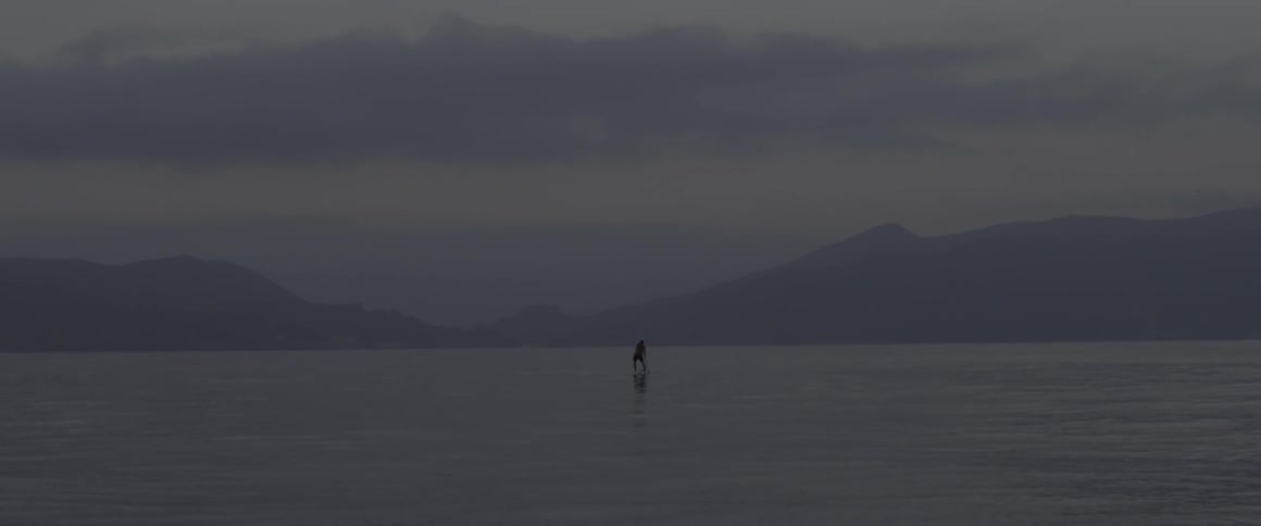 A person is paddling on a board in the distance at sunset