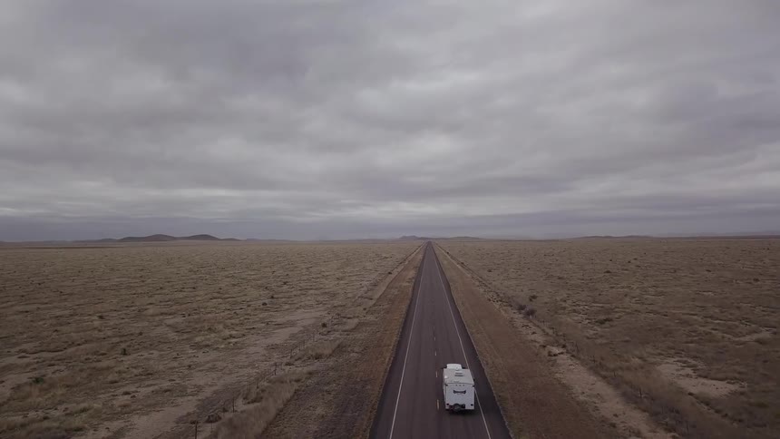 A vehicle towing a trailer drives down a desert road under gray skies
