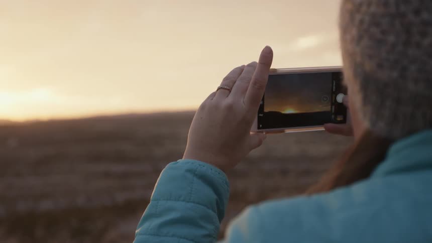Woman standing outside recording video of sunset with camera