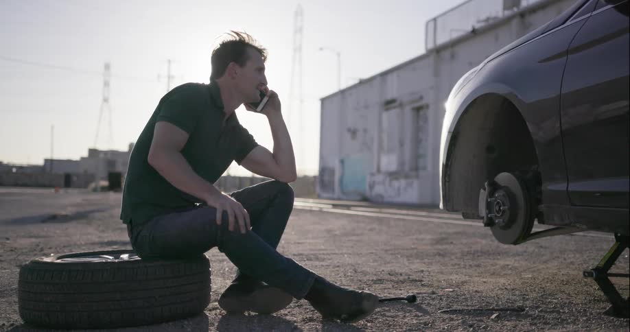 A man sits on a damaged tire talking into cell phone near his vehicle