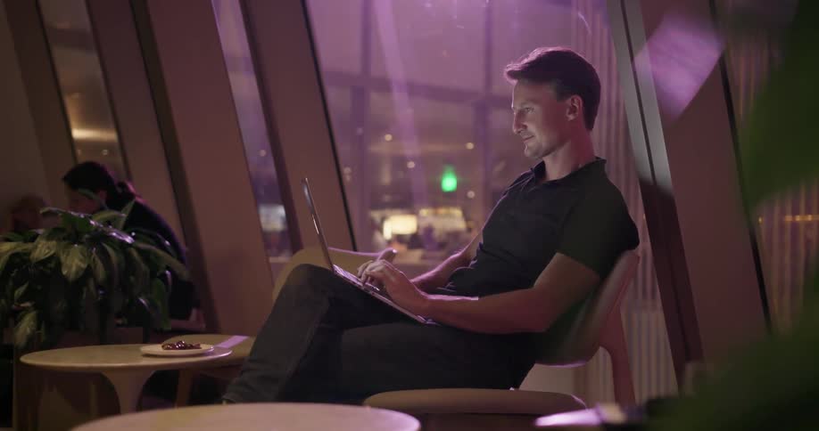A man smiles while typing on a laptop in a public building