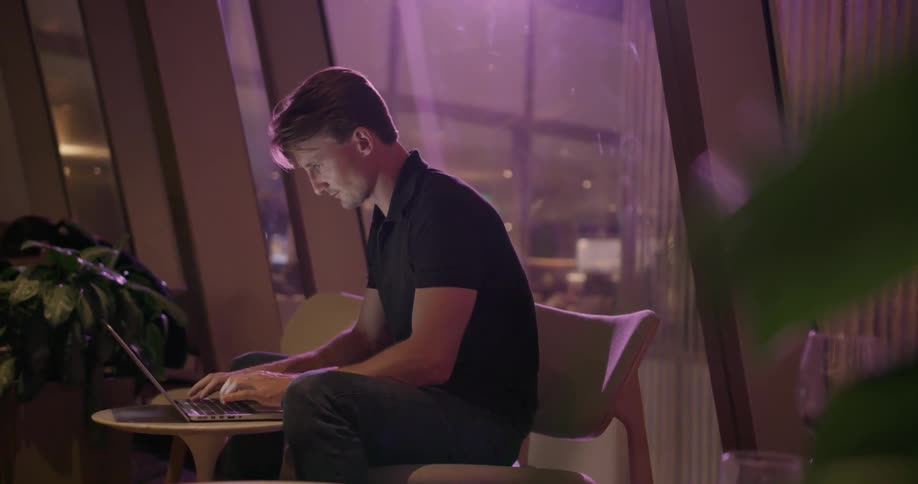 Man sitting inside city building at night typing on laptop