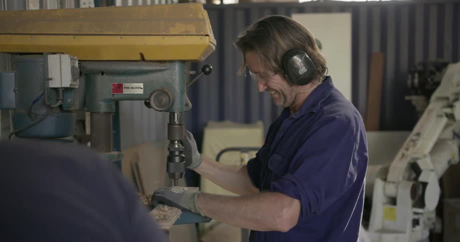 A man smiles and talks to another man as he drills a piece of wood