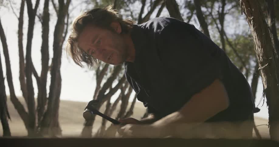 A man chisels a piece of wood outside on a sunny day