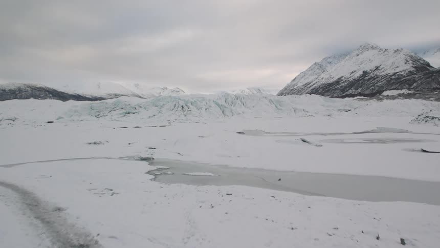 Snow covers rocks and till in front of a glacial landscape with mountains in the distance