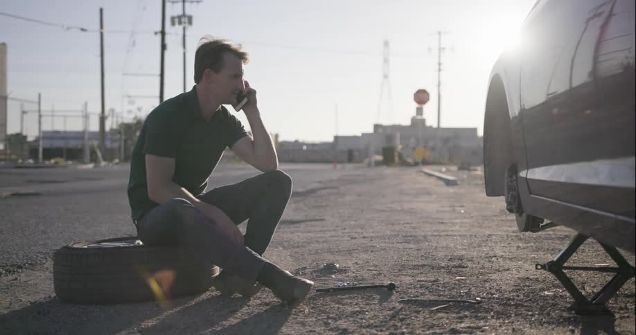 A man sits on a tire and talks on his cell phone