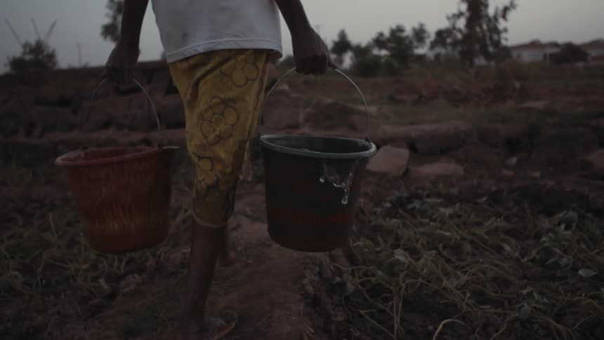 A woman carries buckets of water across a dirt field