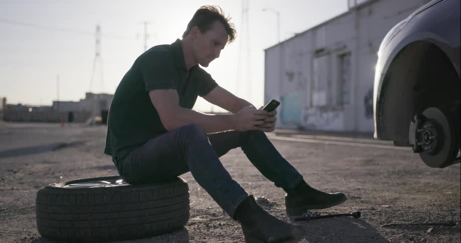 A man sits on a spare tire and types on a smartphone