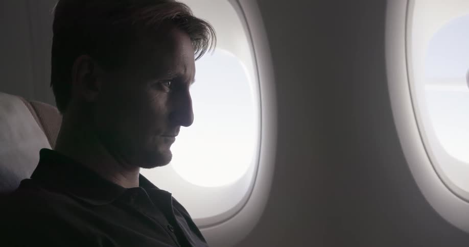 A man sits in an airplane seat typing on his laptop