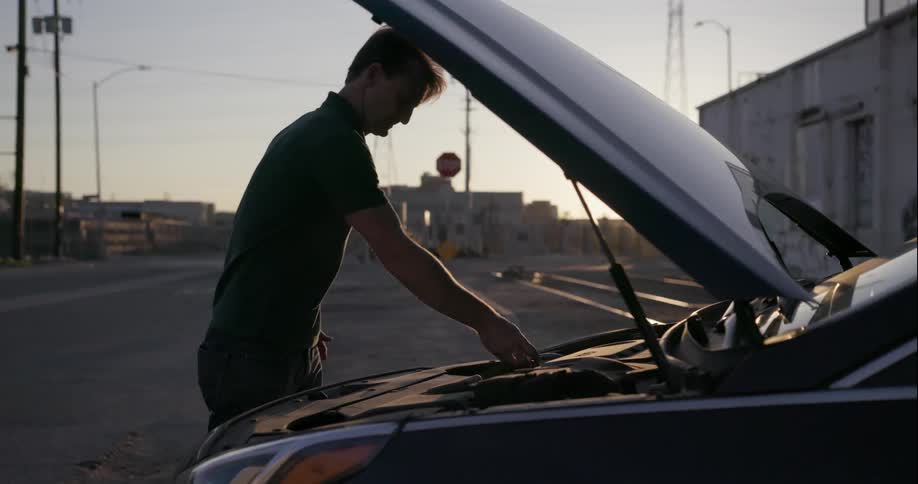 A man looks beneath a car hood while broken down on the side of a road