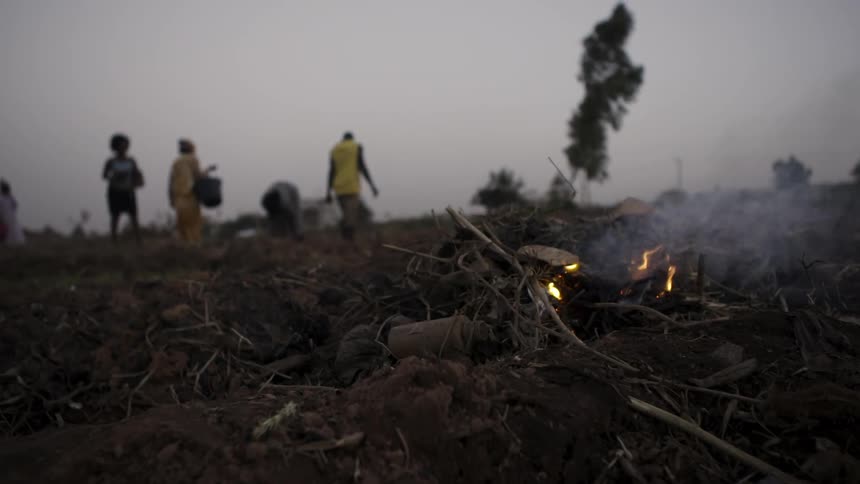 A fire smolders in a rural field with workers in the distance