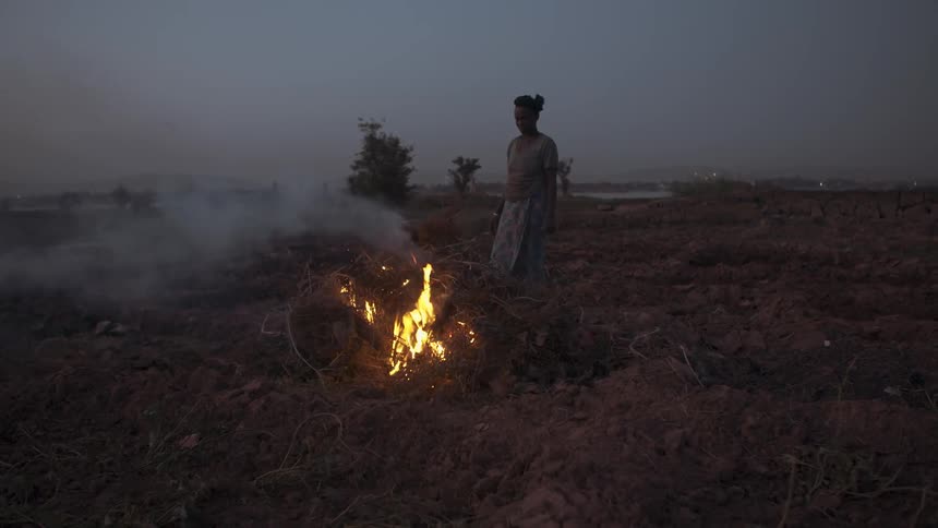 A woman stands near a small campfire at sunset